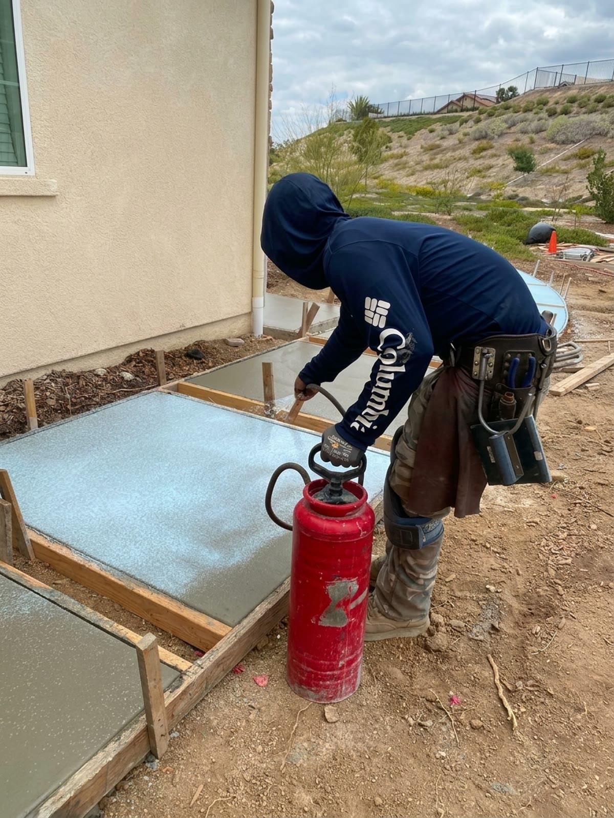 Concrete worker spraying curing compound on a fresh concrete slab beside a home